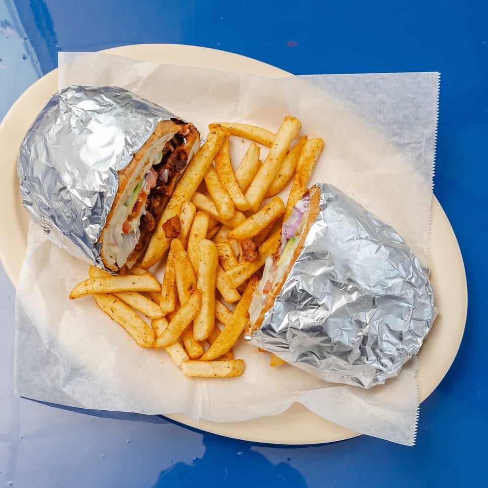 Two foil-wrapped loaded sandwiches with fresh vegetables and seasoned french fries on a paper-lined plate.