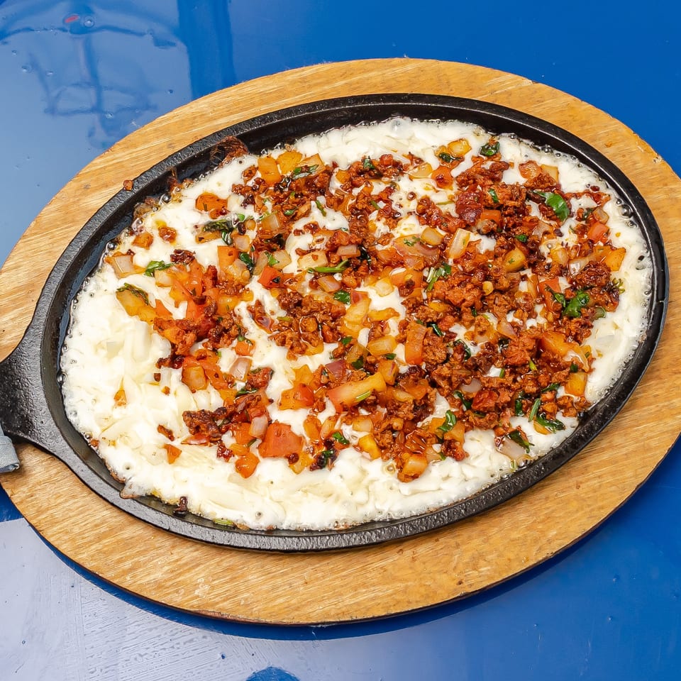 Sizzling skillet filled with melted cheese, diced vegetables, herbs, and browned meat on a wooden serving board.