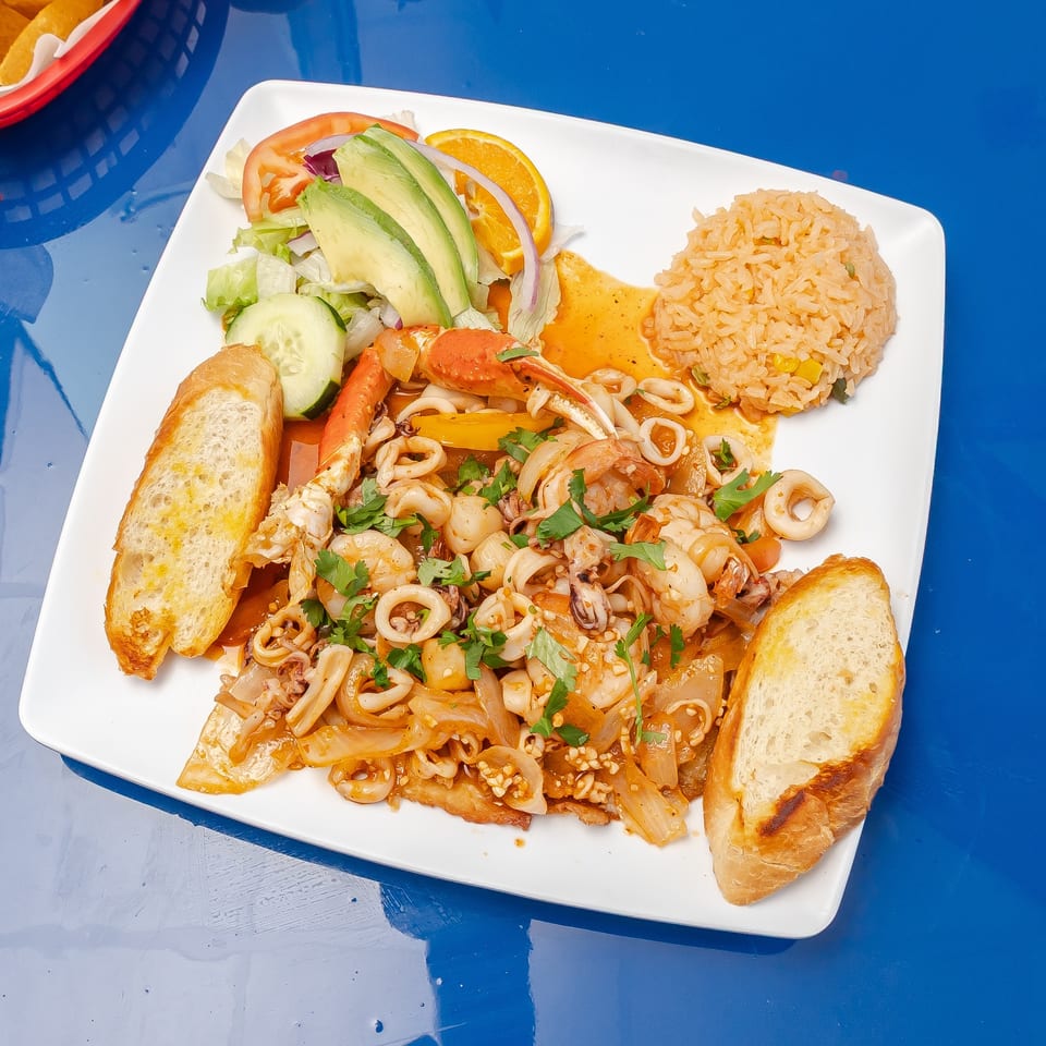 Seafood platter with shrimp, calamari, crab, rice, salad, orange slices, and toasted bread on a white plate.