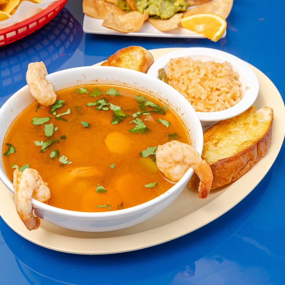 Bowl of shrimp soup garnished with cilantro, served with Mexican rice, toasted bread, tortilla chips, guacamole, and orange wedges.