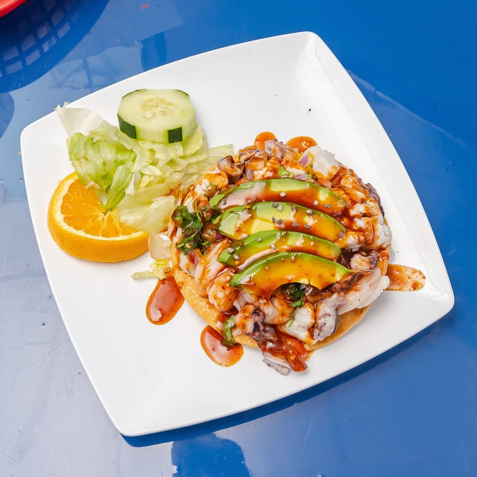 Loaded sandwich topped with avocado, grilled meat, vegetables, and sauce, served with lettuce, cucumber, and orange slices on a white plate.