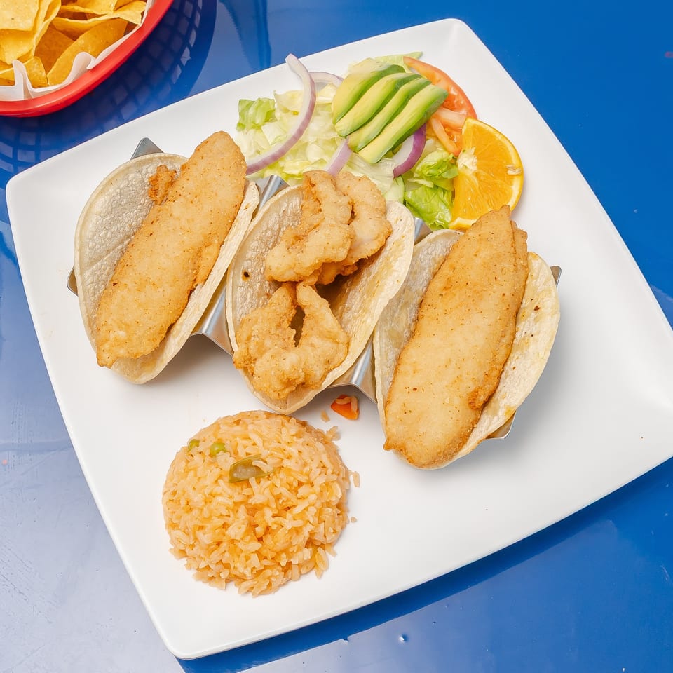 Three crispy fish tacos with avocado salad, orange slices, Mexican rice, and tortilla chips on a white plate.